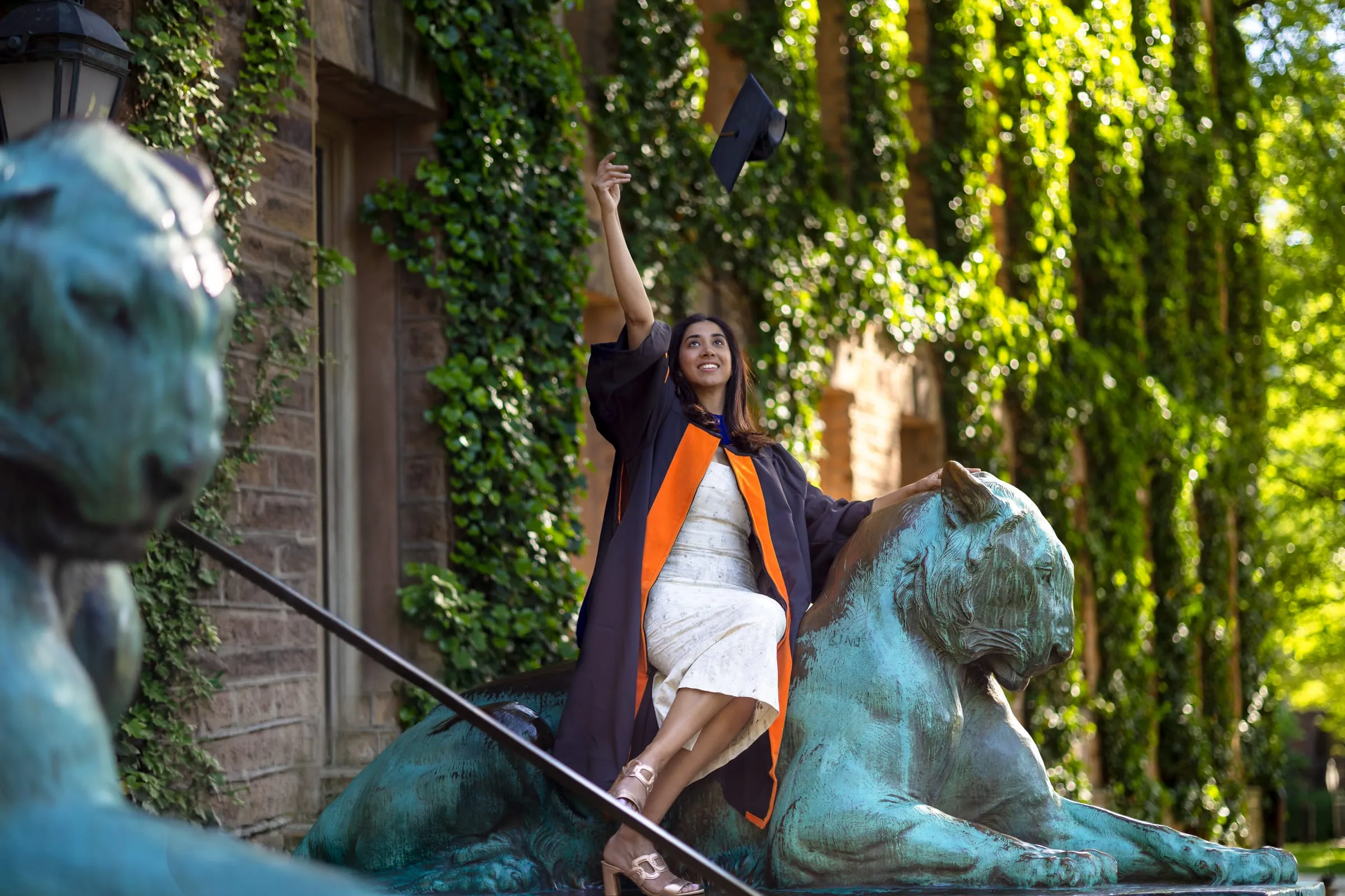 Graduate in a doctoral gown sitting on a bronze tiger statue and raising her cap, ivy-covered walls behind