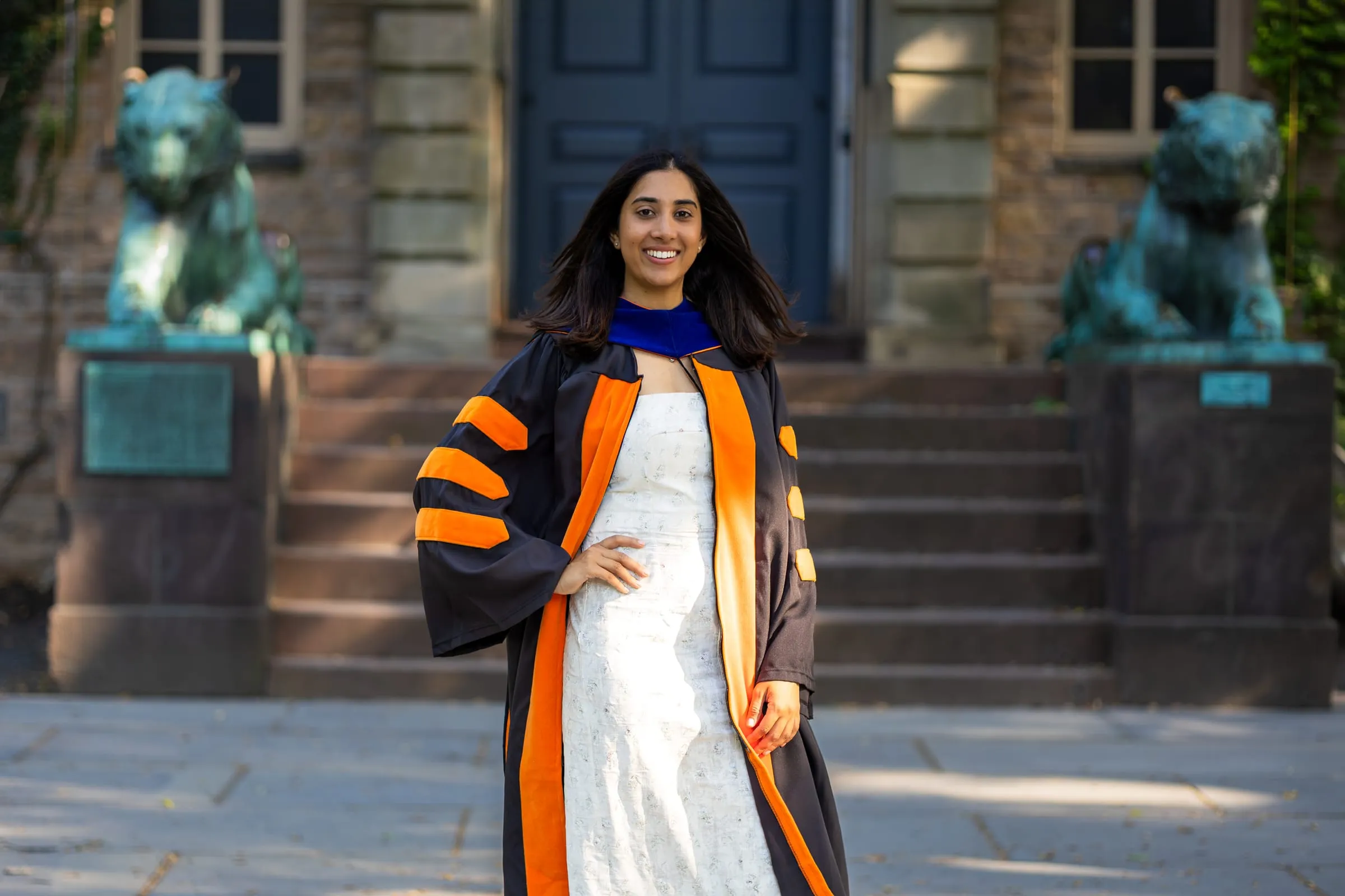 Smiling graduate in an orange and black doctoral gown on stone steps between two bronze tigers