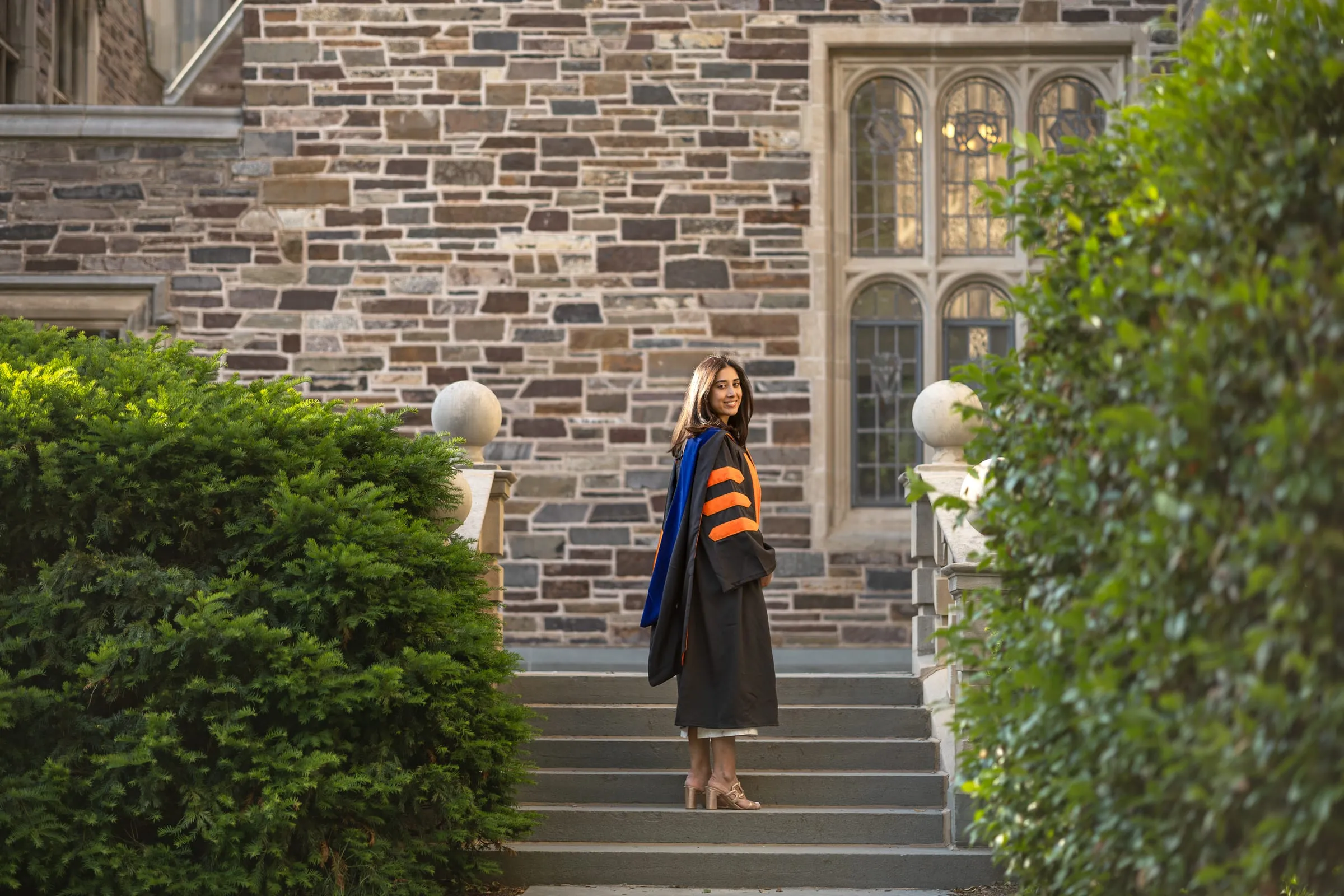 Graduate standing on stone steps between green shrubs and a Gothic building